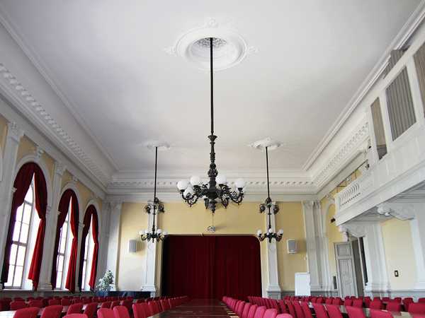 De Geergymnasiets aula med gula väggar, vitt tak med stuckaturer och röda stolar i rader.